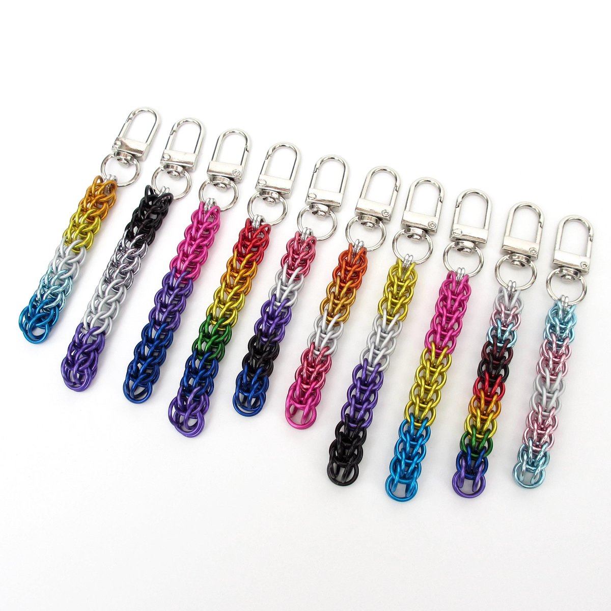 Chainmail Full Persian weave bag charms in ten different Pride flag colors displayed on a white background. Flags shown are AroAce, Asexual, Bisexual, Gay, Genderfluid, Lesbian, Nonbinary, Pansexual, Progress Pride, and Transgender. Each chain hangs from a silver-toned metal swivel clip.